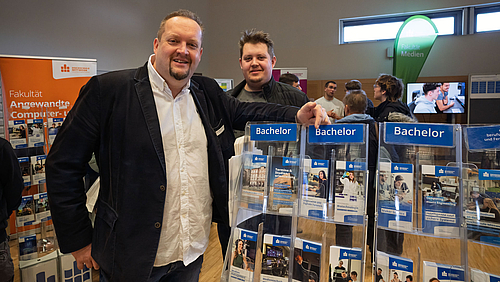 Wie der Vater, so vielleicht der Sohn ... Professor Ronny Bodach empfiehlt Paul den eigenen Studiengang an der HSMW: Allgemeine und Digitale Forensik. Eine ältere und eine jüngere männliche Person stehen lächelnd an einem Ständer mit vielen Studiengangsflyern