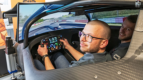 Schulterblick gefahrlos möglich, sonst nah dran am echten Rennerlebnis: Die Maschinenbau-Studenten Max Walter und Philipp Goldammer (v.l.) in der Large Scale Simulationplattform des „KTM xBow GT4“-Rennwagens im Labor der Hochschule Mittweida. Zwei junge Männer im Cockpit eines Rennwagens, das Teil einer Simulationsplattform ist, in der das Umfeld Rennstrecke auf großen Monitoren dargestellt ist. Der linke Mann fasst mit beiden Händen das Lenkrad. Beide schauen nach hinten in Richtung Kamera.
