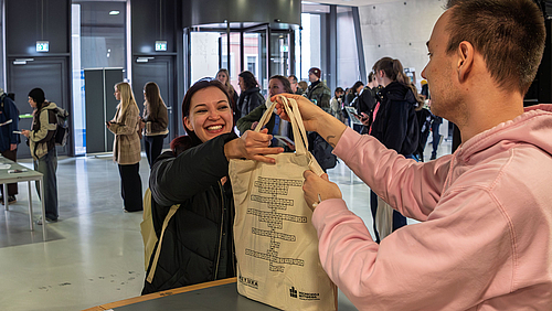Das Studium in der Tasche – jedenfalls die Erstausstattung aus den Händen des Studierendenrats inklusive Mensagutschein, Kochlöffel, Energiedrink und vielem mehr. Eine strahlend lächelnde weibliche Person erhält aus den Händen einer männlichen Person einen Stoffbeute über einen Tresen gereicht.