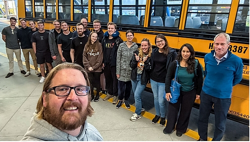 Mittweida goes Rochester. Professor Martin Anselm vom RIT begrüßt die frisch gelandete Gruppe der HSMW. Selfie zeigt eine Gruppe von überwiegend jungen Personen vor einem typischen gelben us-amerikanischen Schulbus. Im Bildvordergrund ist der Kopf des Fotograf zu sehen.